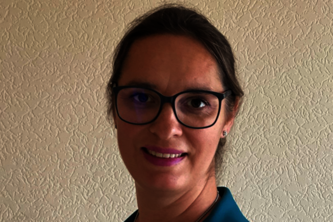 Anika Schlaud Frau mit Brille und zurückgebundenem Haar trägt ein türkisfarbenes Sakko und lächelt vor einer hellen, strukturierten Wand