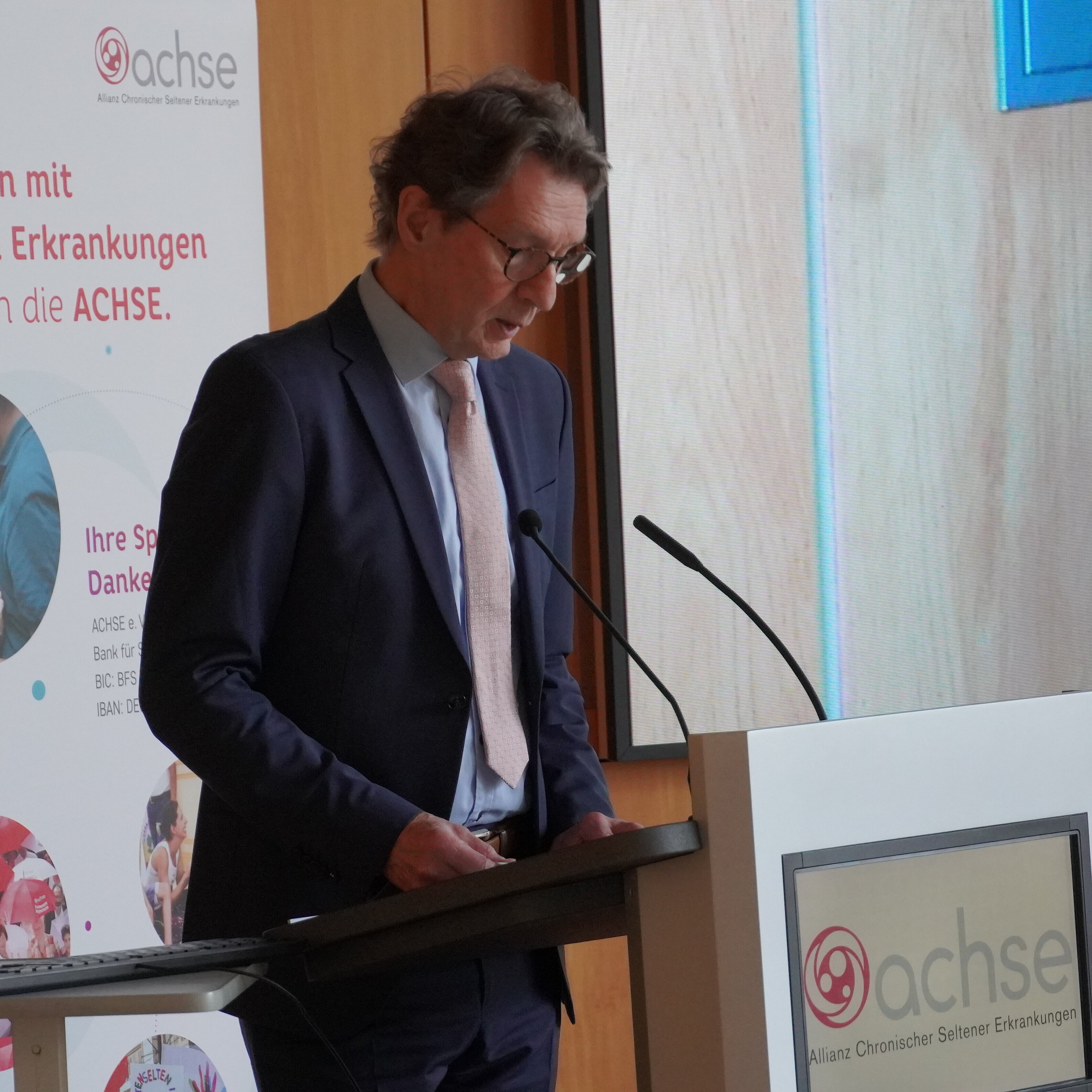 Mann im Anzug steht an einem Rednerpult mit Mikrofon, im Hintergrund Roll-up mit Text und Logo der ACHSE.