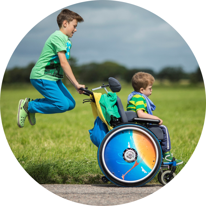 Ein Junge schiebt einen anderen Jungen, der im Rollstuhl sitzt. Dabei stützt sich der schiebende Junge an den Griffen in die Höhe. Beide Kinder tragen grüne, farbenfrohe Oberteile. Der Rollstuhl hat ein buntes Rad mit einem Farbverlauf. Im Hintergrund sind unscharf eine Wiese und dahinter Bäume zu erkennen. 