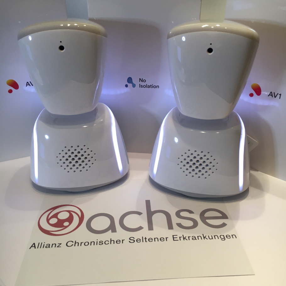 Zwei weiße Avatare mit Lautsprechern unten und kleinen Kameralinsen oben stehen nebeneinander auf einem Tisch mit dem ACHSE Logo.