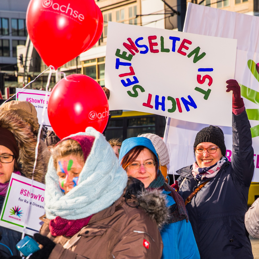 Menschen bei einer Demonstration halten bunte Schilder und rote Luftballons mit dem Schriftzug 'achse'. Ein Schild zeigt den Text 'SELTEN IST NICHT SELTEN' in bunten Buchstaben.