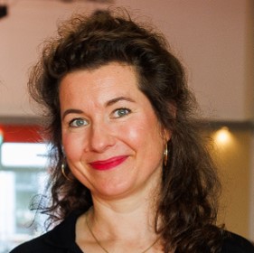 Bianca Paslak-Leptien, mit lockigem, schulterlangem Haar, trägt eine schwarze Bluse und goldene Ohrringe. Der Hintergrund ist unscharf.