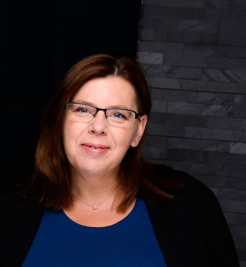 Porträt einer Frau mit mittellangem braunen Haar, die eine Brille trägt. Sie hat ein blaues Oberteil und eine schwarze Jacke an und steht vor einem dunklen, strukturierten Hintergrund.