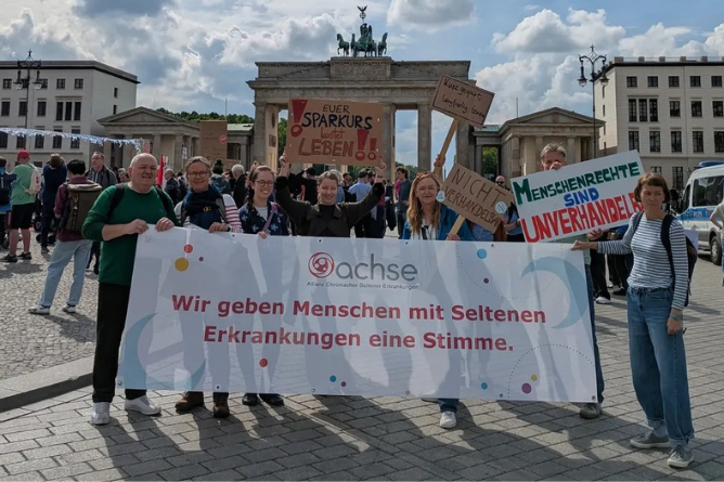 Gruppe von Menschen vor dem Brandenburger Tor hält Banner mit der Aufschrift 'Wir geben Menschen mit Seltenen Erkrankungen eine Stimme' und weitere Schilder mit Forderungen zu Menschenrechten und Lebenssparplänen