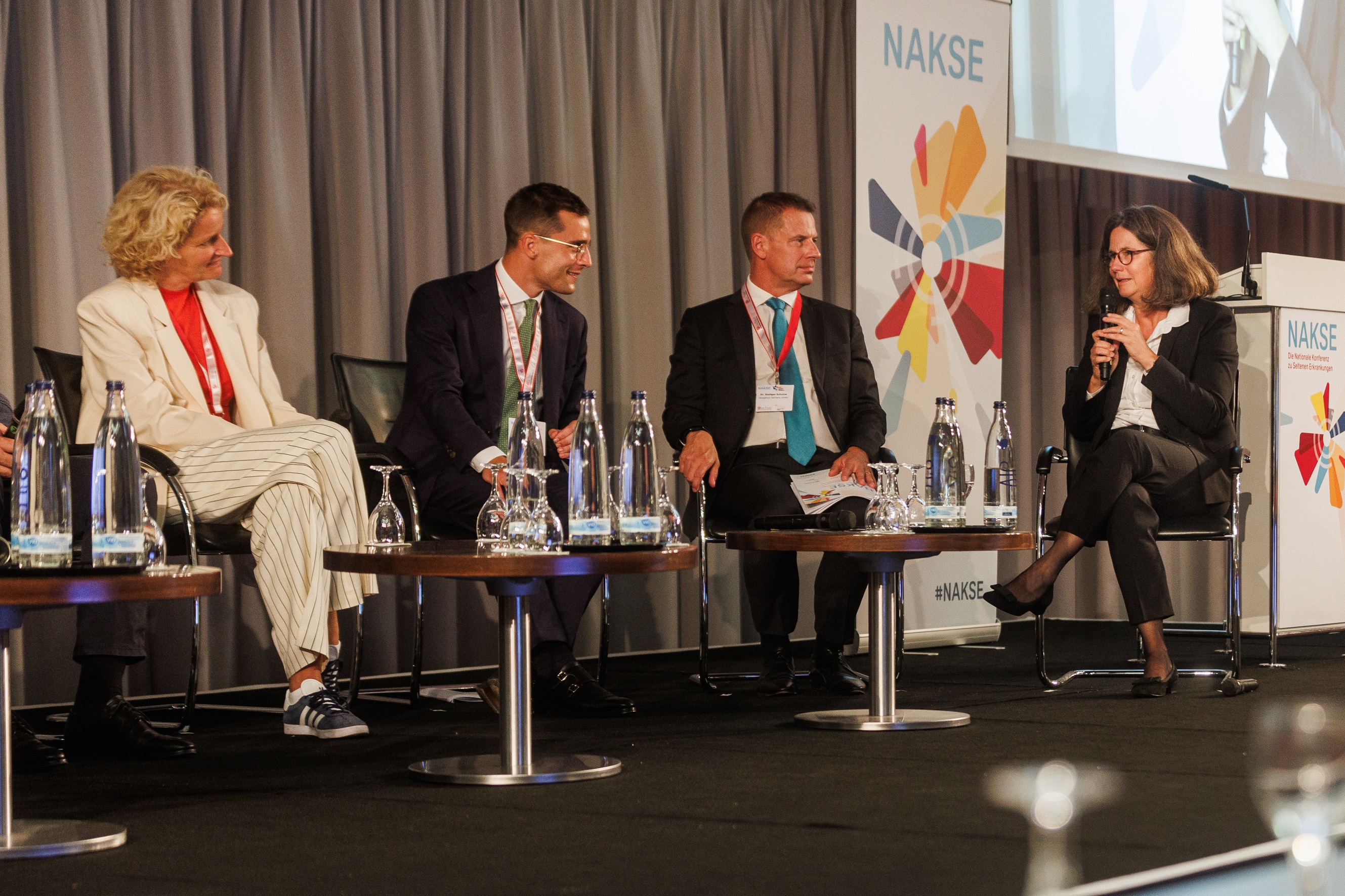Zwei Frauen, zwei Männer im Halbkreis diskutieren im Rahmen der NAKSE. Vor ihnen, auf kleinen ruinden Tischen, stehen Wasserflaschen und Gläser. Mirjam Mann sitzt rechts außen und spricht in ein Mikrofon