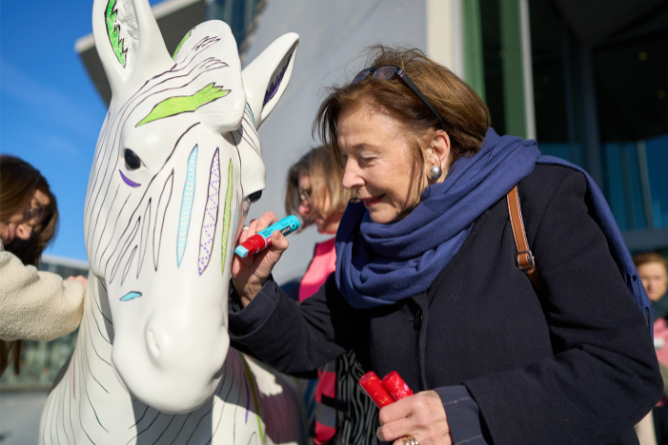 Frau mit blauem Schal bemalt eine weiße Zebra-Skulptur mit bunten Markern im Freien