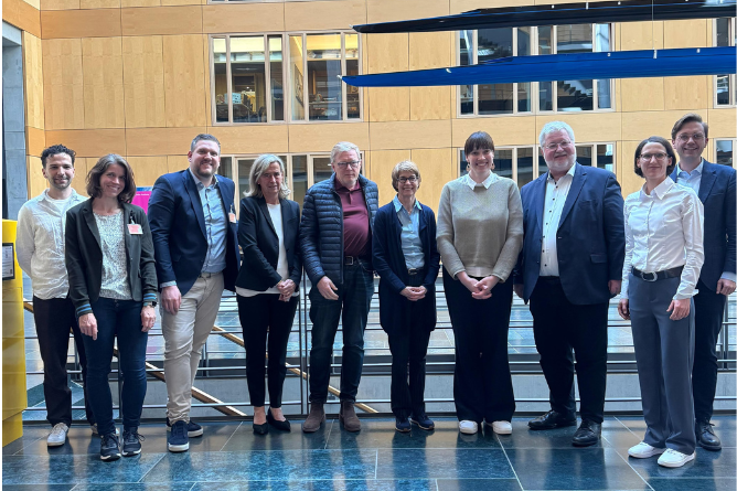 Gruppe von neun Personen steht in einem modernen Innenraum vor einer Glasbrüstung und einer Holzpaneelwand