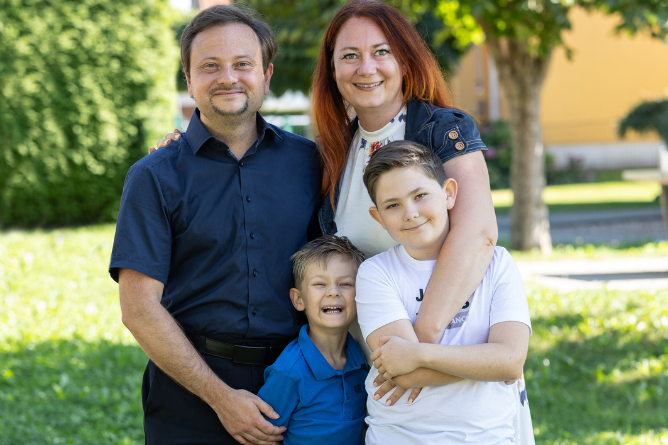 Familie mit zwei Kindern steht eng umarmt im Freien vor einem Baum und einem gelben Haus