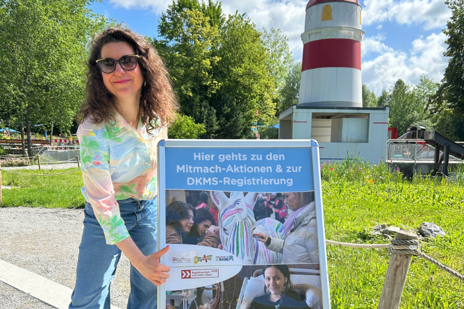 © Ravensburger Spieleland 2026 Person mit lockigem Haar hält ein Schild mit der Aufschrift 'Hier gehts zu den Mitmach-Aktionen & zur DKMS-Registrierung' vor einem Leuchtturm im Freien