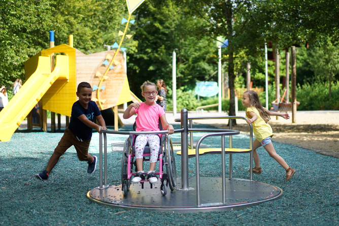 Drei Kinder spielen auf einem Karussell auf einem Spielplatz, eines sitzt im Rollstuhl, die anderen halten sich fest und laufen mit