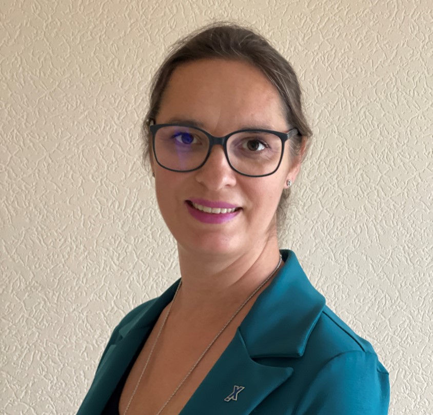 Eine Frau mit dunkel gerahmter Brille, nach hinten gebundenen dunkelbraunen Haaren, einem petrolfarbenen Oberteil und einer Halsette 