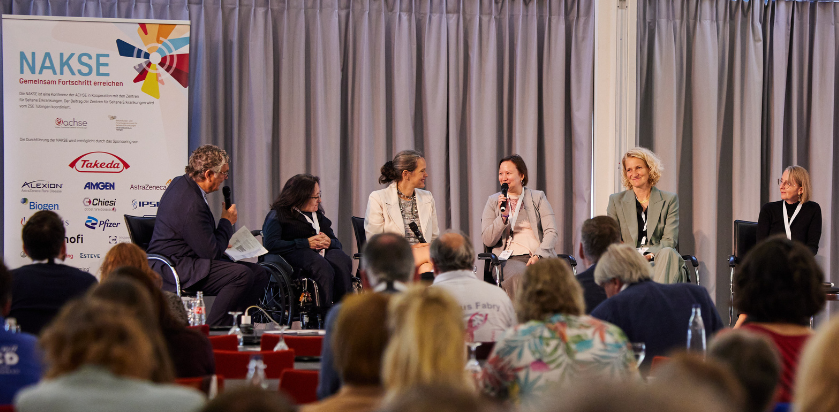 Podiumsdiskussion mit fünf Personen auf einer Bühne vor Publikum, links ein Banner mit dem Schriftzug 'NAKSE' und mehreren Logos von Unternehmen und Organisationen.
