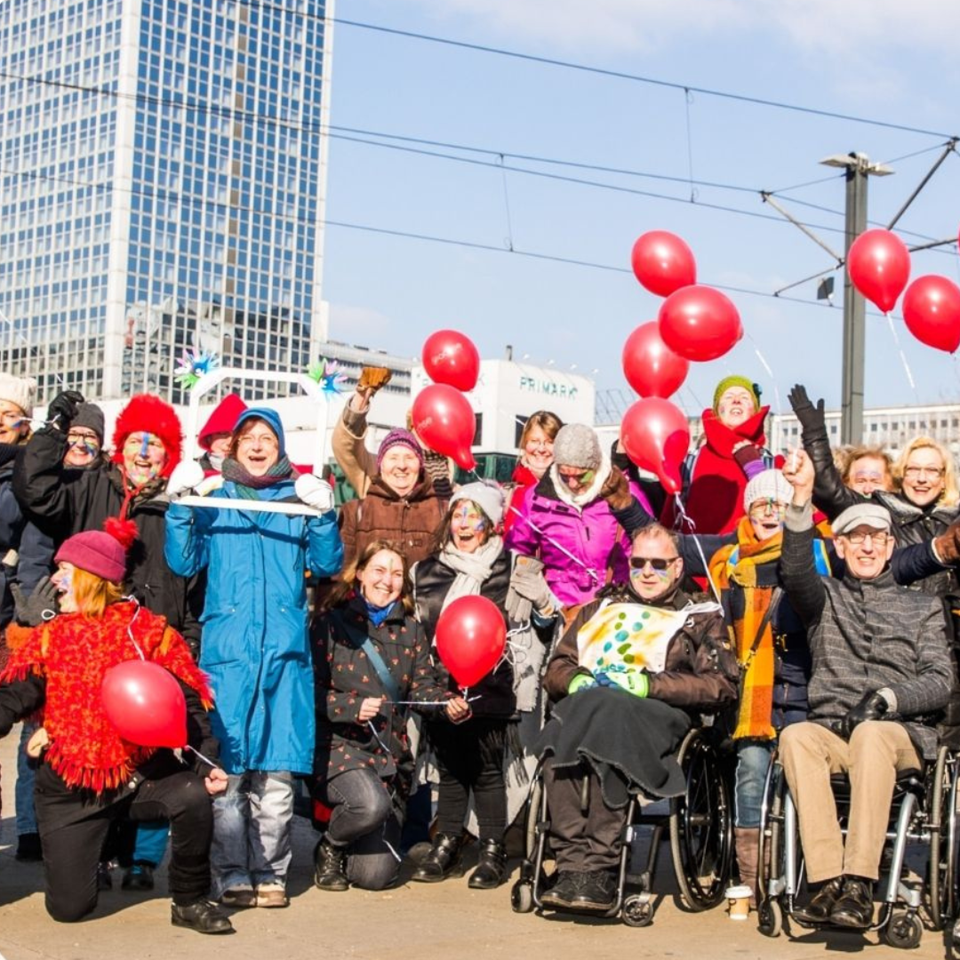 Gruppe von Menschen mit roten Luftballons, darunter eine Person im Rollstuhl, vor einem Hochhaus im Freien