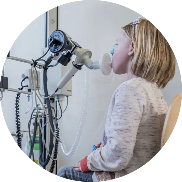 Ein blondes Kind sitzt an einem medizinischen Gerät zur Lungenfunktionsprüfung, Mundstück vor dem Gesicht, trägt hellen Pullover