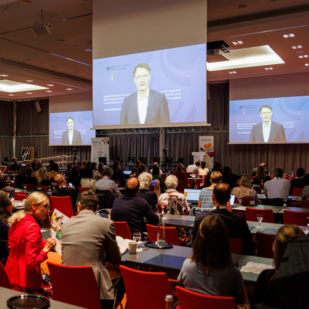 Konferenzsaal mit Publikum vor Bühne und drei großen Leinwänden, die Bundesgesundheitsminister Karl Lauterbach bei seinem Redebeitrag zeigen