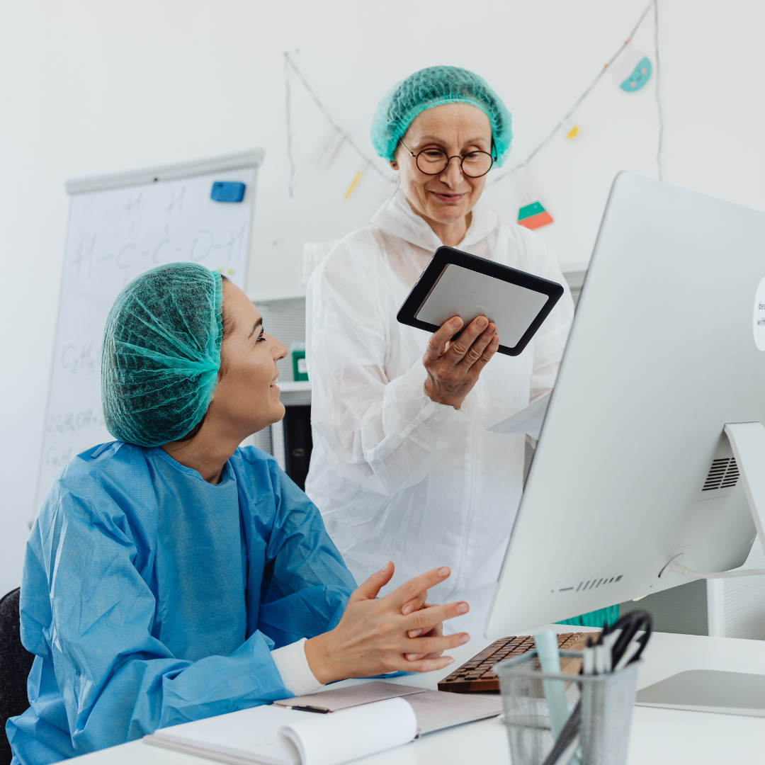 Zwei Personen in medizinischer Schutzkleidung und Haarnetzen vor einem Computer, eine hält ein Tablet