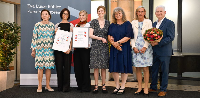 Gruppe von sieben Personen in formeller Kleidung, zwei halten Urkunden, eine hält einen Blumenstrauß