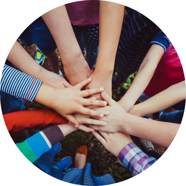 Ein Foto von mehreren Händen, die sich in der Mitte stapeln, als Symbol für Gemeinschaft und Zusammenarbeit. Die Personen tragen bunte Kleidung, darunter gestreifte und karierte Muster. Der Hintergrund zeigt Gras und vereinzelt Schuhe, die zu den stehenden Personen gehören.