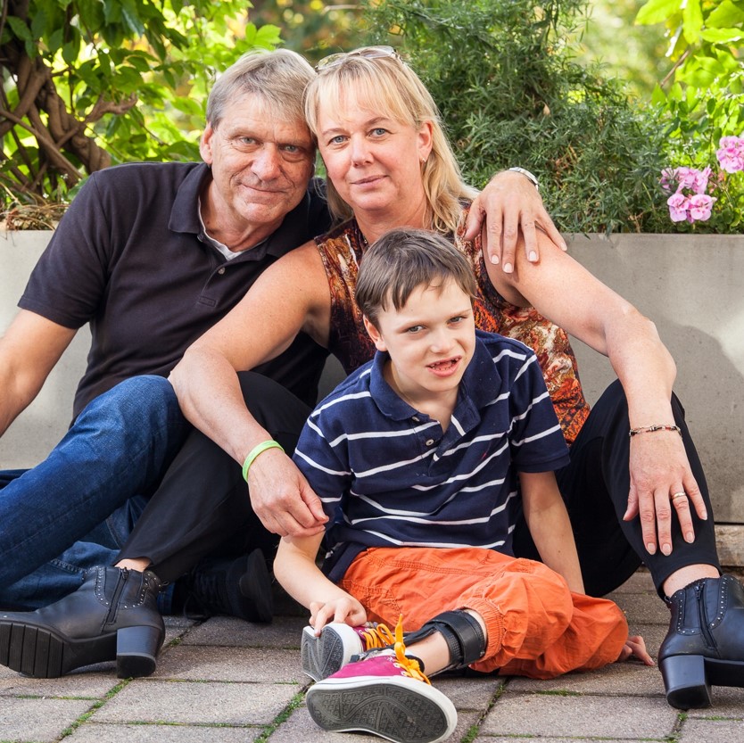 Drei Personen sitzen zusammen vor einer bepflanzten Mauer, ein Mann und eine Frau umarmen ein Kind mit Beinprothese, Gründer der Selbsthilfegemeinschaft Hand in Hand gegen Tay-Sachs und Sandhoff e.V.