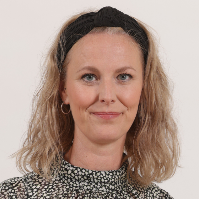 Carina Thomas Frau mit schulterlangem, gewelltem Haar und gemustertem Oberteil vor hellem Hintergrund