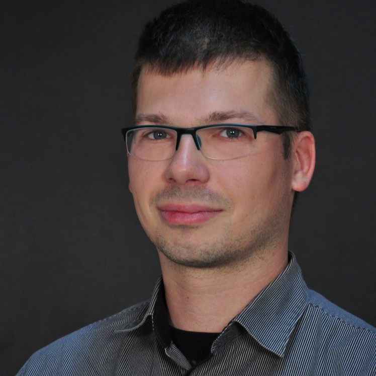 Mann mit kurzem dunklem Haar, trägt eine gestreifte Hemdjacke über schwarzem T-Shirt, vor dunklem Hintergrund