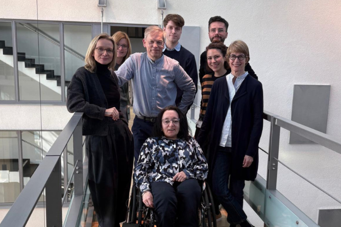 Gruppe von sechs Personen, darunter eine im Rollstuhl, steht auf einem Balkon mit Glasgeländer in einem modernen Innenraum