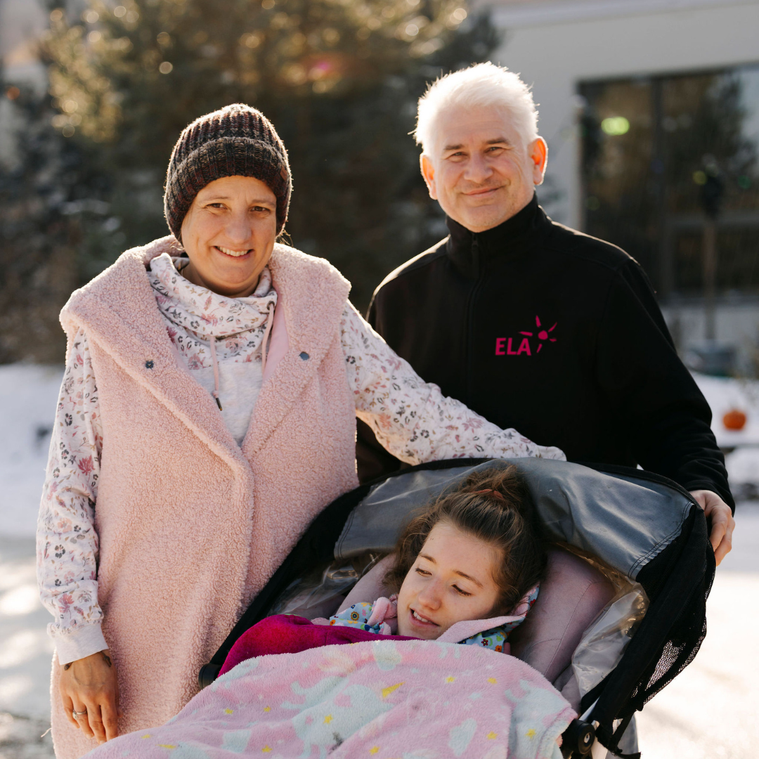 Drei Personen im Freien, eine Person sitzt in einem Kinderwagen mit einer Decke, die anderen beiden stehen daneben, eine trägt eine rosa Weste, die andere ein schwarzes Oberteil mit rotem Schriftzug