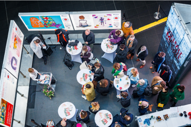 Menschen stehen und sitzen an Stehtischen in einer Ausstellungshalle mit bunten Kunstwerken an der Wand