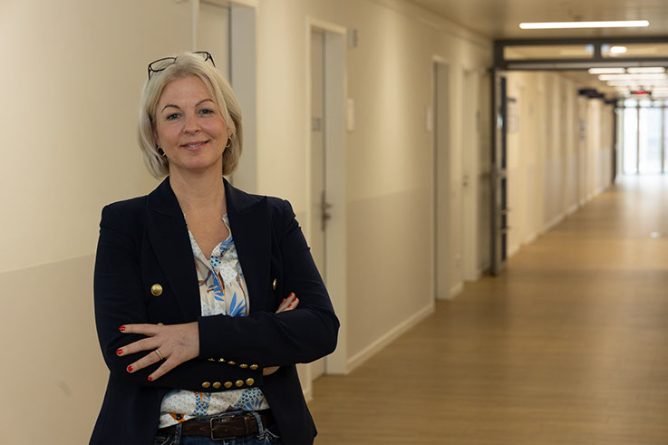 Frau mit verschränkten Armen, dunklem Blazer mit goldfarbenen Knöpfen und gemustertem Hemd steht in einem langen, hellen Flur mit mehreren Türen