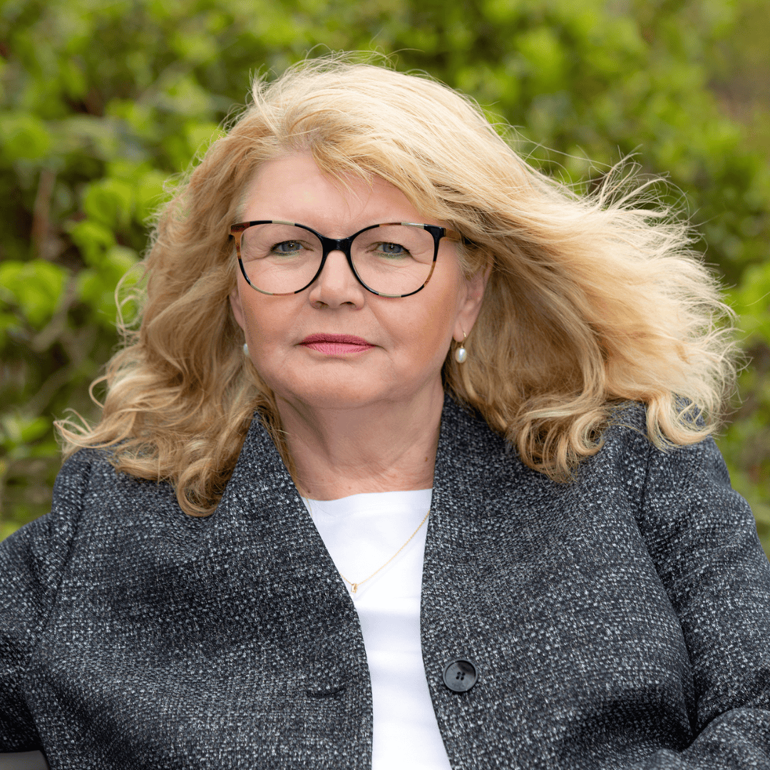 Frau mit langen, lockigen blonden Haaren trägt eine dunkelgraue Jacke und ein weißes Oberteil vor grünem Hintergrund