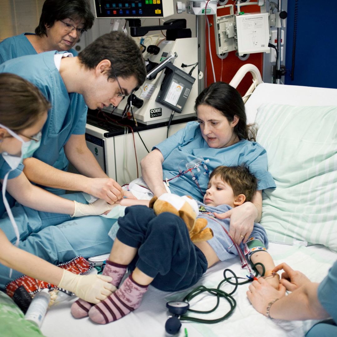 Mehrere Pflegekräfte betreuen ein Kind im Krankenhausbett, das ein Kuscheltier hält und an medizinische Geräte angeschlossen ist.
