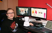 Person sitzt an einem Schreibtisch mit zwei Monitoren, einer Tastatur, einer weißen Roboterfigur und einem Laptop vor roter Wand.