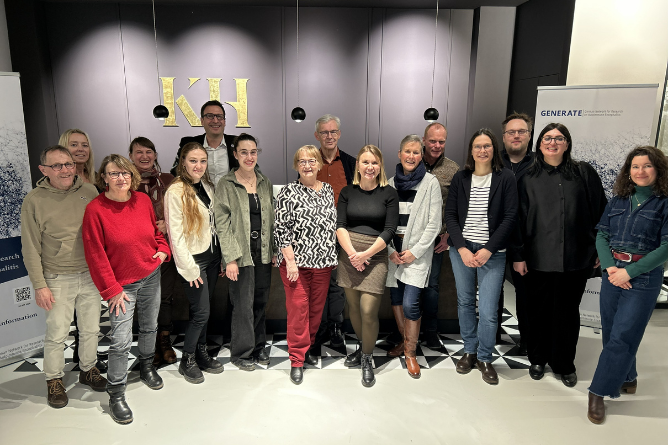 Gruppenfoto von Menschen in einem Raum mit dunkler Wand und goldenen Buchstaben KH im Hintergrund