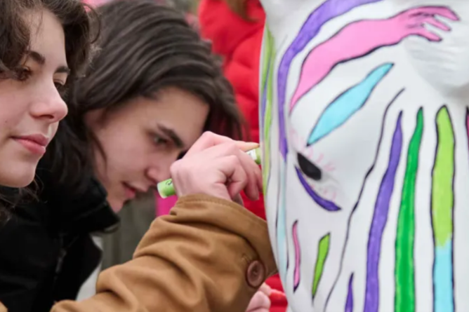 Junge Frau mit dunklen Locken und brauner Jacke malt bunte Streifen auf eine weiße Maske