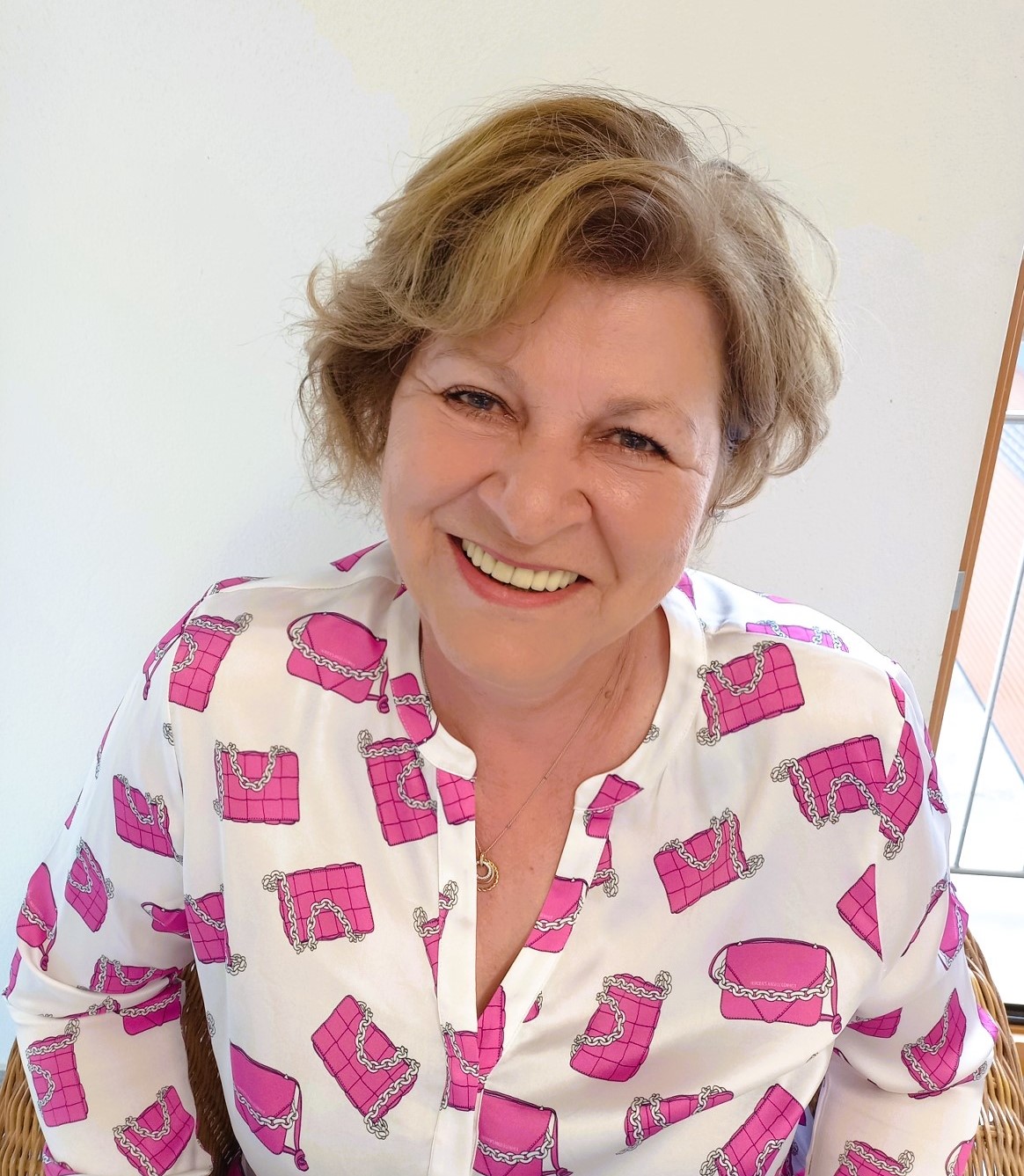 Eine Frau mit blondem, halblangem Haar sitzt auf einem Stuhl vor einer weißen Wand. Sie trägt eine weiße Bluse mit auffälligem Muster aus pinkfarbenen Handtaschen mit Kettendetails. Im Hintergrund ist ein Fenster mit hellem Tageslicht zu sehen. Eine Frau mit blondem, halblangem Haar sitzt auf einem Stuhl vor einer weißen Wand. Sie trägt eine weiße Bluse mit auffälligem Muster aus pinkfarbenen Handtaschen mit Kettendetails. Im Hintergrund ist ein Fenster mit hellem Tageslicht zu sehen.