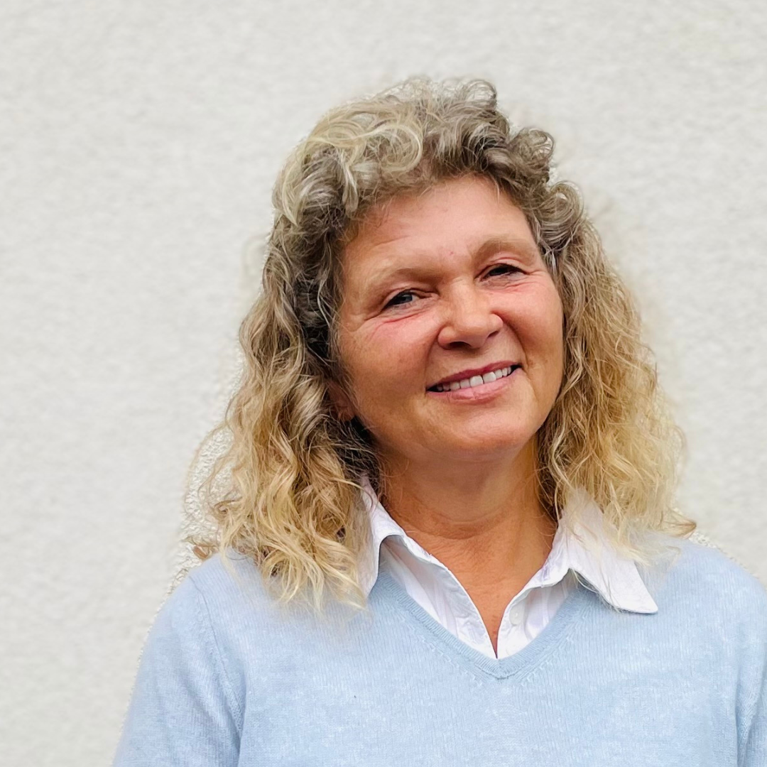 Frau mit lockigem Haar, hellblauen Pullover über weißem Hemd vor einer hell verputzten Wand