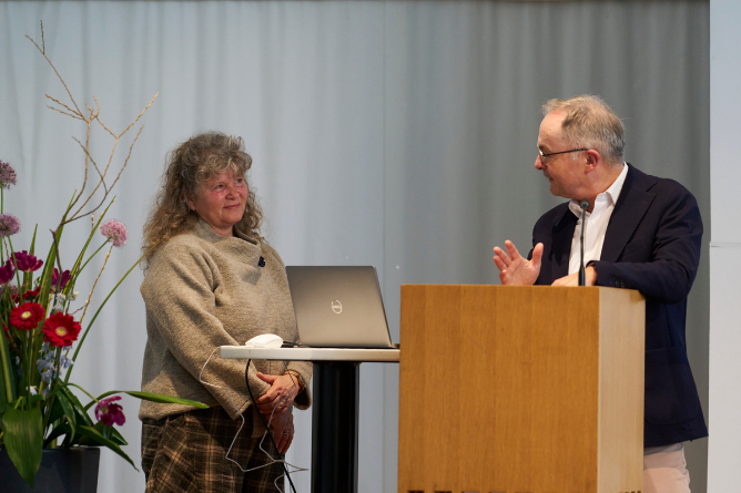Zwei Personen stehen an einem Rednerpult und einem kleinen Tisch mit Laptop vor einem hellen Vorhang, links davon ein großer Blumenstrauß