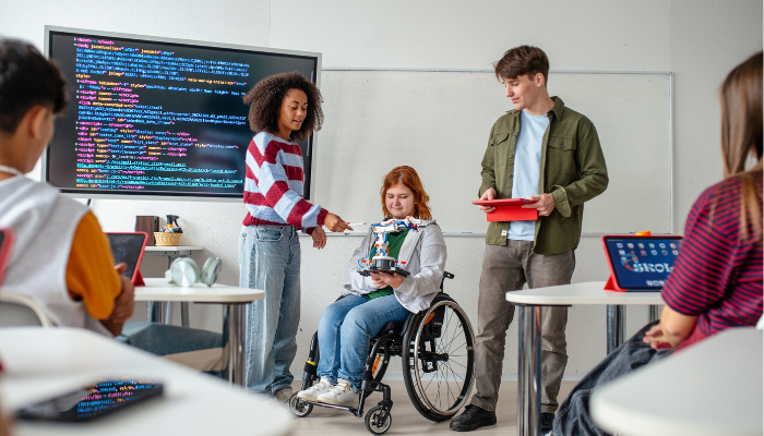 Drei junge Menschen, eine im Rollstuhl, zeigen in einem Klassenraum mit Computern und großem Bildschirm mit Programmiercode eine technische Konstruktion