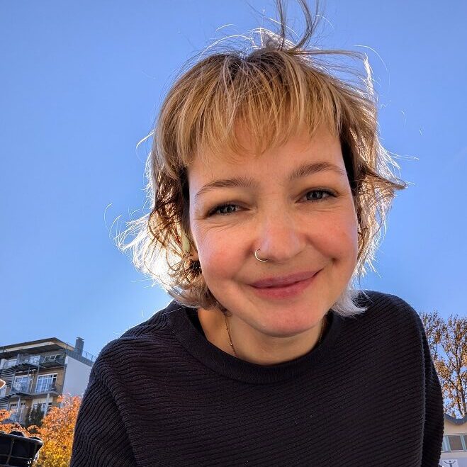 Person mit schulterlangem, gewelltem, blondem Haar trägt schwarzen Pullover, im Freien vor blauem Himmel und Gebäuden.