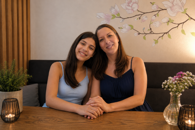 Zwei Frauen sitzen eng nebeneinander an einem Holztisch, halten sich an den Händen und lächeln in die Kamera
