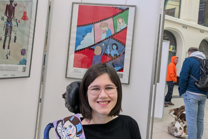 Person mit schwarzem Oberteil sitzt vor einer Wand mit zwei gerahmten Bildern, eines zeigt eine Comicfilmstreifen-ähnliche Illustration mit roten und blauen Flächen