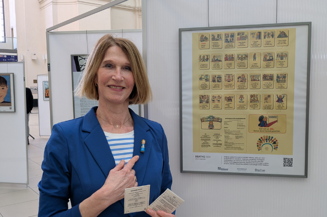 Person mit schwarzem Oberteil sitzt vor einer Wand mit zwei gerahmten Bildern, eines zeigt eine Comicfilmstreifen-ähnliche Illustration mit roten und blauen Flächen