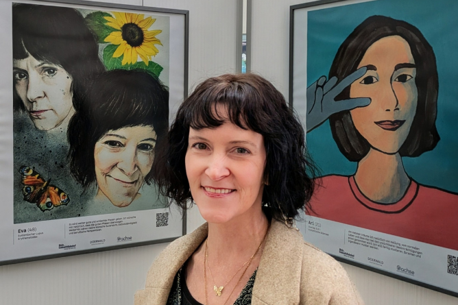 Frau mit schulterlangem Haar steht vor zwei farbigen Gemälden, eines zeigt eine Sonnenblume und einen Schmetterling, das andere eine stilisierte Person mit erhobener Hand.