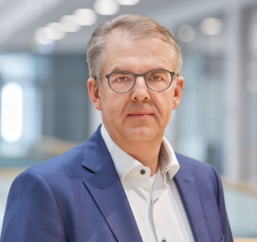 Mittelalter Mann mit grauen, anch hinten gekämmten Haaren, grau gefasster Brille mit fast runden Gläsern, weißem Hemd und blauem Sakko