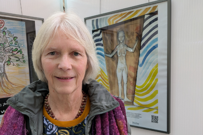 Ältere Person mit weißem Haar, bunter Kleidung und Perlenkette vor einem farbigen Gemälde mit einer menschlichen Figur und wellenförmigen Mustern an der Wand