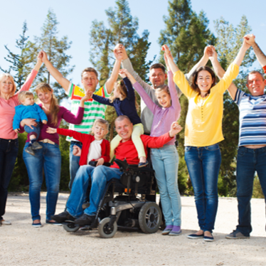 Eine Gruppe von Menschen unterschiedlichen Alters steht draußen auf einem sonnigen Platz vor Bäumen. Alle halten sich an den Händen und heben die Arme freudig in die Luft. In der Mitte sitzt ein Mann im Rollstuhl, auch er ist Teil der fröhlichen Runde. Kinder und Erwachsene sind dabei. Die Stimmung wirkt freundlich und gemeinschaftlich.