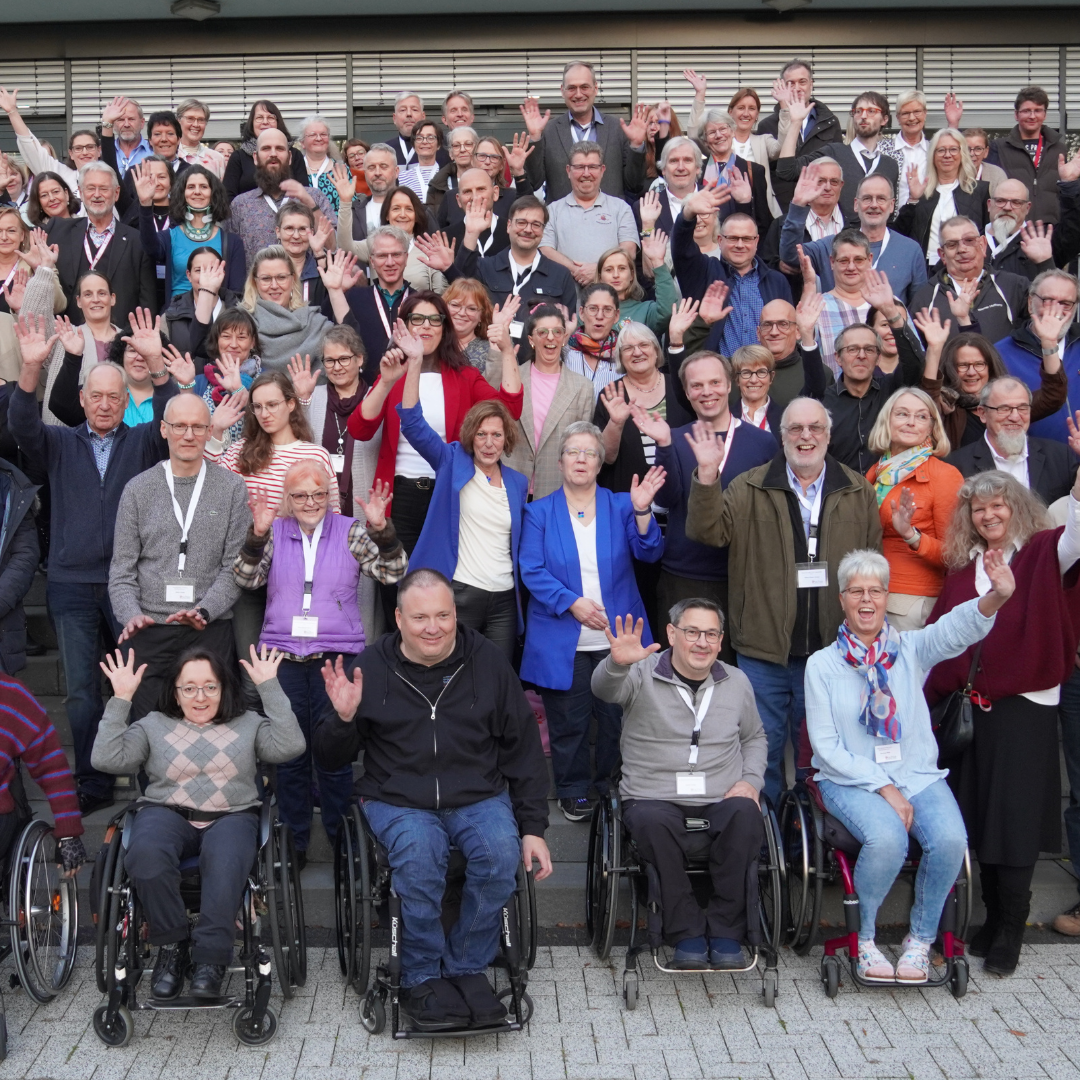 Große Gruppe von Menschen, darunter mehrere Personen im Rollstuhl, posiert vor einem mehrstöckigen Gebäude auf einer Treppe und winkt in die Kamera.