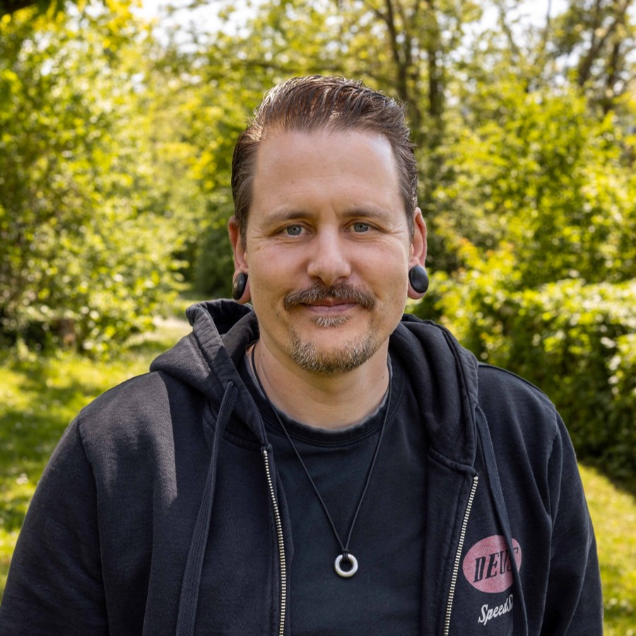 Martin Armbruster, Selbsthilfegemeinschaft CdLS, lächelnd im Freien mit schwarzer Kapuzenjacke und schwarzem T-Shirt, trägt schwarzen Ohrplug und Halskette Martin Armbruster, Selbsthilfegemeinschaft CdLS, lächelnd im Freien mit schwarzer Kapuzenjacke und schwarzem T-Shirt, trägt schwarzen Ohrplug und Halskette