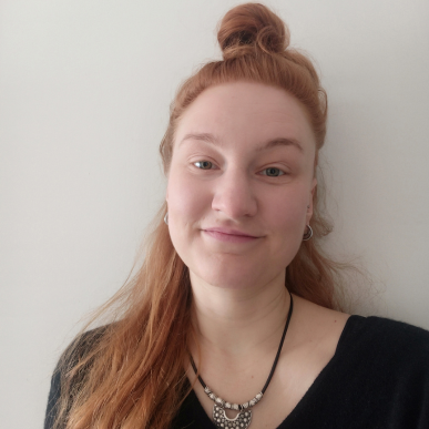 Lillie Kannegießer Frau mit langem roten Haar, das zu einem Dutt hochgesteckt ist, trägt schwarzen Pullover und eine Halskette mit schwarzem Band vor weißer Wand
