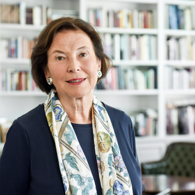 Porträt von Eva Luise Köhler, der Schirmherrin der ACHSE e. V., mit kinnlangem, braunem Haar. Sie trägt ein dunkelblaues Oberteil und einen farbenfrohen Schal mit Blumenmuster. Sie steht in einem Raum vor einem Bücherregal und lächelt leicht in die Kamera.